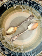 Two Sterling Silver ornate spoons on a textured ceramic plate with decorative patterns.
