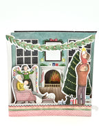A 3D holiday greeting card depicting a warm Christmas scene with a decorated tree and gifts, a fireplace, and people enjoying the holiday. The card has a 'Merry Christmas' greeting on the back.