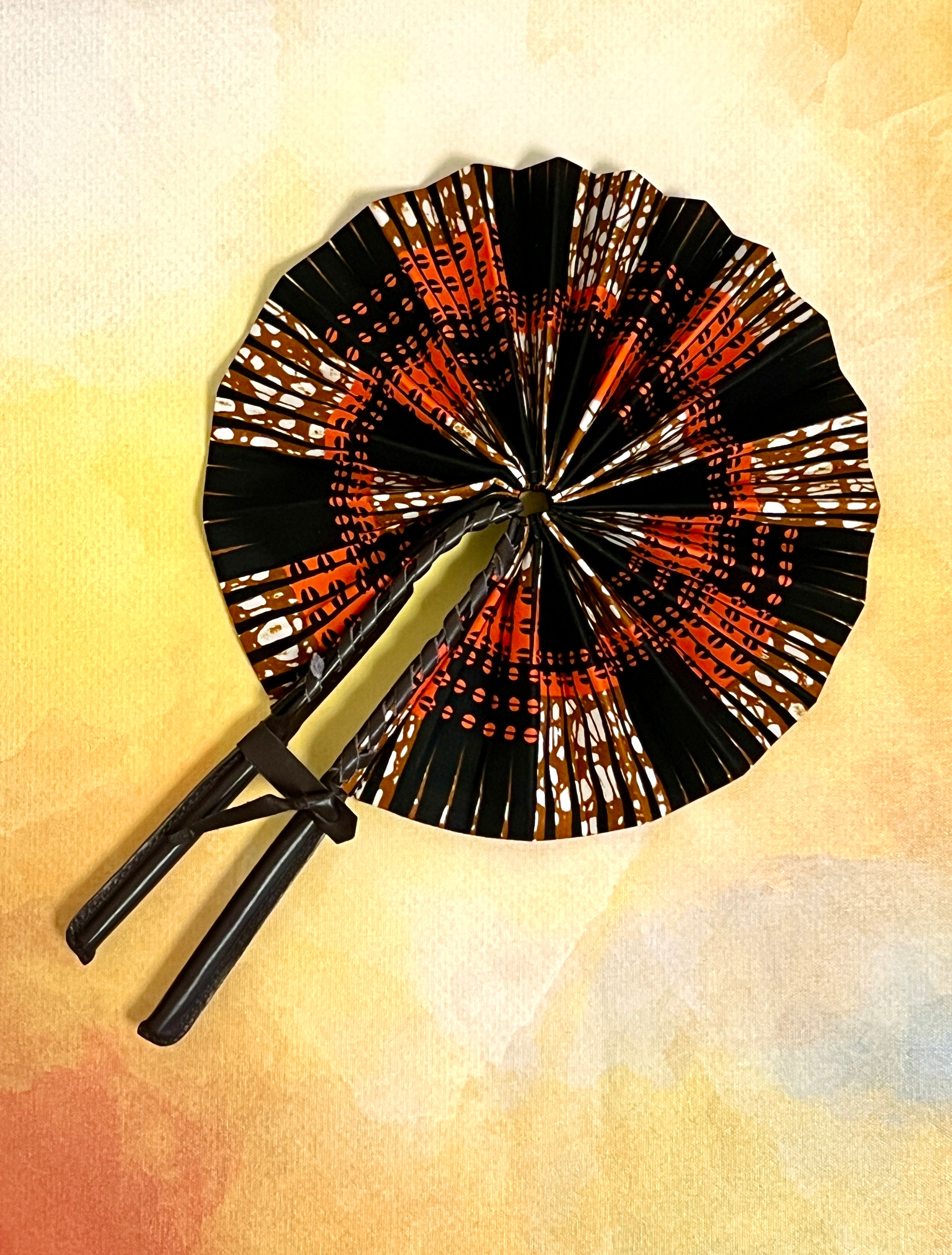 An open Ulla Johnson hand fan with Ankara wax print design, featuring a leather handle and closure, displayed against an abstract background.